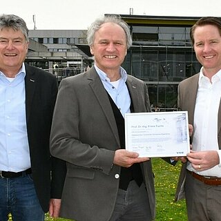 Drei Männer stehen vor einem Gebäude. Zwei Männer halten gemeinsam eine Urkunde.