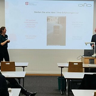 Blick in einen Schulungsraum mit Leinwand. An einer Leinwand referieren 3 Männer und eine Frau.
